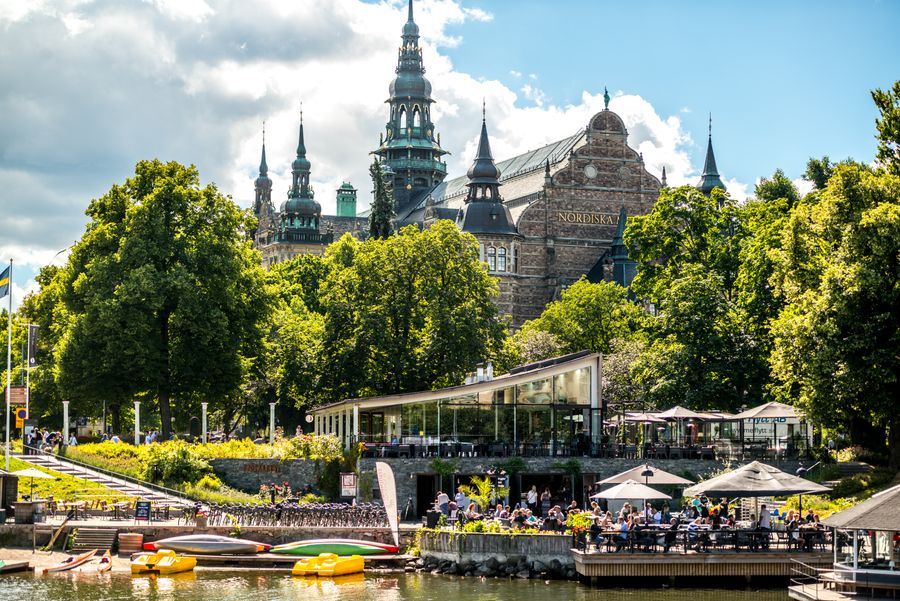  Et bilde av Nordiska museet i Stockholm med en café i forgrunnen, tatt fra vannkanten. 