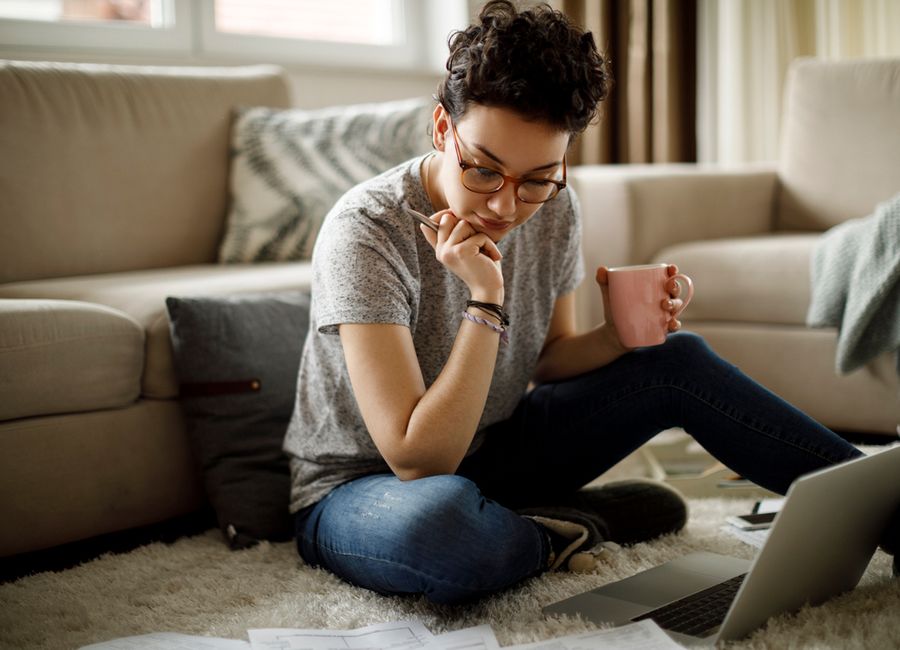 En kvinne som sitter på gulvet foran laptopen sin mens hun holder en penn i den ene hånden og en kaffekopp i den andre.