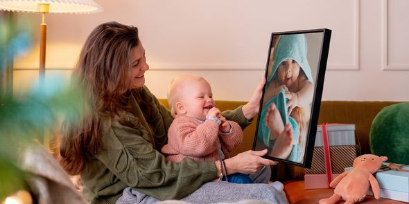 Kvinne med en baby på fanget i en stue som holder opp personlig veggkunst av babyen, mens babyen ser på den og fniser, som illustrerer fotoknudsen sin guide med fotogaver til babyens første jul.