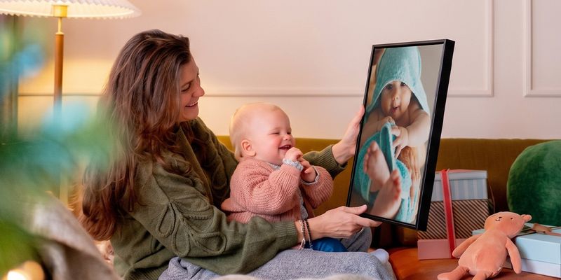 Kvinne med en baby på fanget i en stue som holder opp personlig veggkunst av babyen, mens babyen ser på den og fniser, som illustrerer fotoknudsen sin guide med fotogaver til babyens første jul.