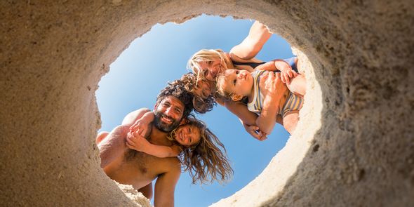 En familie på fire på stranden – mamma, pappa, datter og en liten gutt – ser ned i et hull gravd i sanden, og bildet fanger et lekent sommerøyeblikk som inspirerer til strandbilder og strandfotografering.