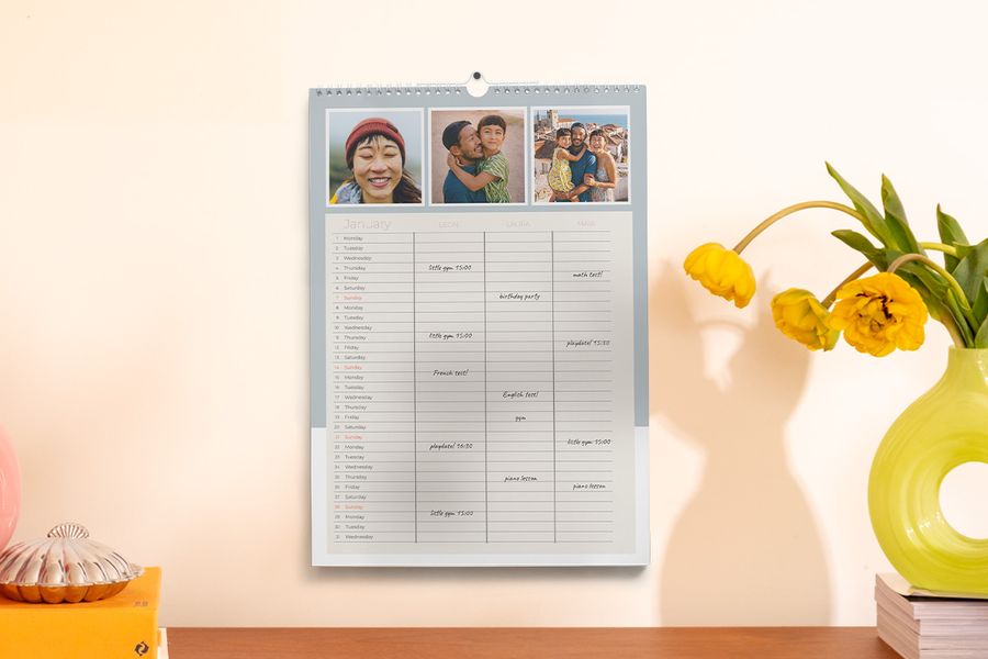 En familieplanlegger henger på veggen ved siden av en vase og noen bøker, som illustrerer fotoknudsen sitt tips om å holde seg organisert gjennom skoleåret med en personlig planlegger.