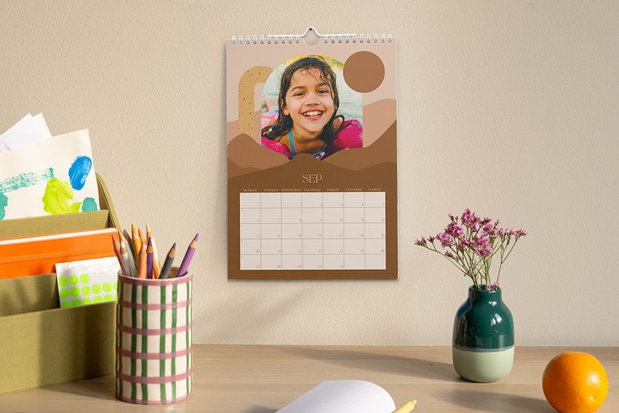 En personlig kalender som viser september og et bilde av en ung jente, henger over et skrivebord med en pennebeholder, en vase med blomster og en oppbevaringsboks med kunst- og skolesaker, som illustrerer fotoknudsen sitt tips om å skape en fokusert arbeidsplass for skoleåret.