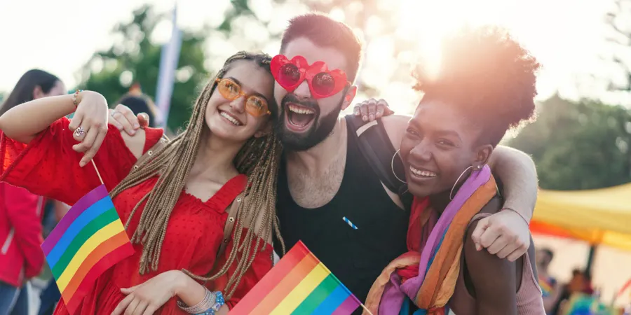 Tre venner smiler på et Pride-arrangement, holder regnbueflagg og har på seg solbriller, og feirer gleden og samholdet i LGBTQI+ Pride.