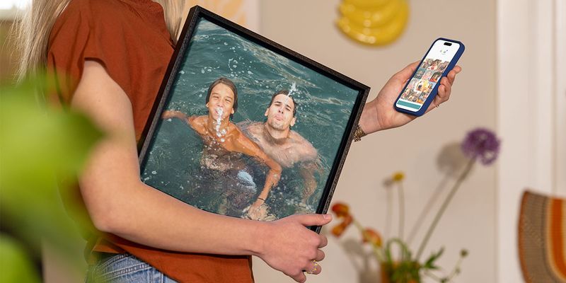 En kvinne holder et innrammet feriebilde av en mann og en liten gutt som leker i vannet under armen, mens hun velger ut flere bilder på telefonen sin, som illustrerer fotoknudsen sin guide til å gjøre feriebilder om til vegg-kunst.