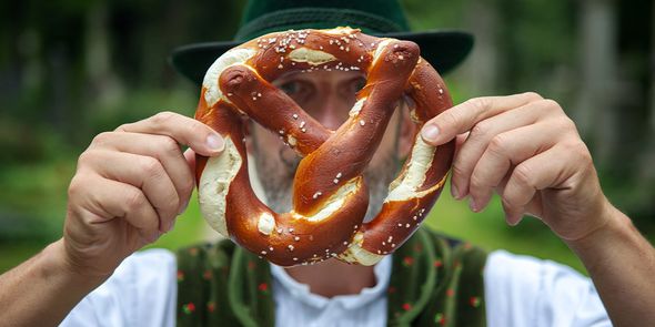 En mann i tradisjonell bayersk klesdrakt holder en stor pretzel foran ansiktet, som introduserer fotoknudsen sin guide til de beste Oktoberfest-byene i Europa.