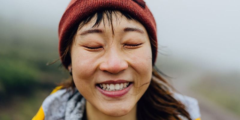 Smilende kvinne med lukkede øyne utendørs i vinterklær og rød lue, som illustrerer fotoknudsen sine 75 ideer til bildetekster for høstbilder på Instagram.