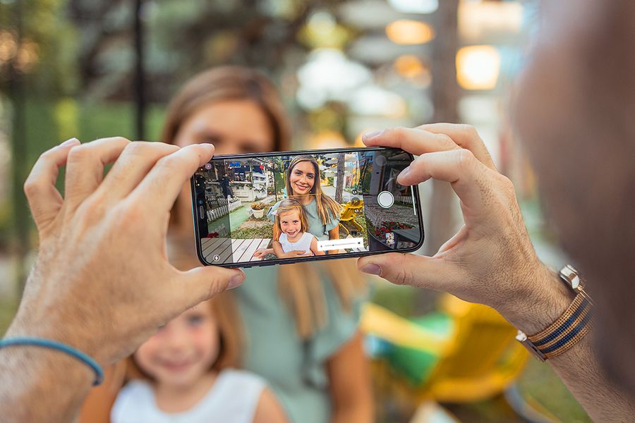 En hånd holder en mobiltelefon som tar bilde av en smilende mor og datteren hennes i sterkt sollys, og illustrerer hvordan du kan ta bilder i sterkt sollys med mobilen.