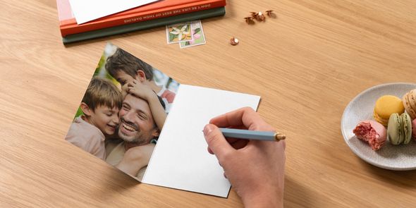 Et åpent personlig fotoknudsen farsdagskort viser en far med sine to sønner, med en penn klar til å skrive en hilsen ved siden av noen bøker og en tallerken med makroner, som inspirasjon til tekst til farsdagskort.