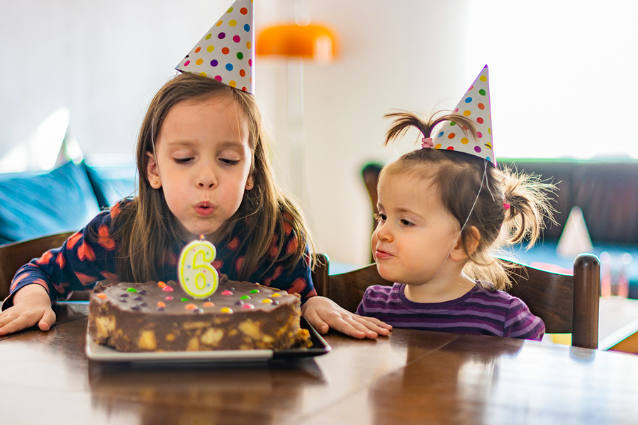 En liten jente med festhatt blåser ut lyset på kaken til 6-årsdagen sammen med en venn, som viser personlige bursdagsgaver til barn fra fotoknudsen.