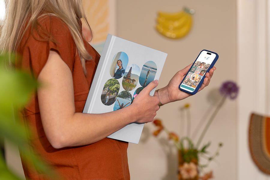 En kvinne holder et lukket fotoknudsen feriealbum med familiebilder fra reisen på forsiden, samtidig som hun ser på reisebilder på mobilen, og illustrerer hvordan man velger de riktige bildene til en personlig fotobok reise.