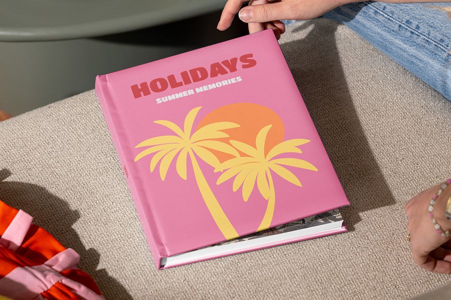 En lukket fotoknudsen fotobok reise med tittelen «Holidays, summer memories» og illustrerte palmer på et rosa omslag holdes av hender som er klare til å åpne den, som inspirasjon til hvordan du kan strukturere et feriealbum og fortelle reisefortellingen din.