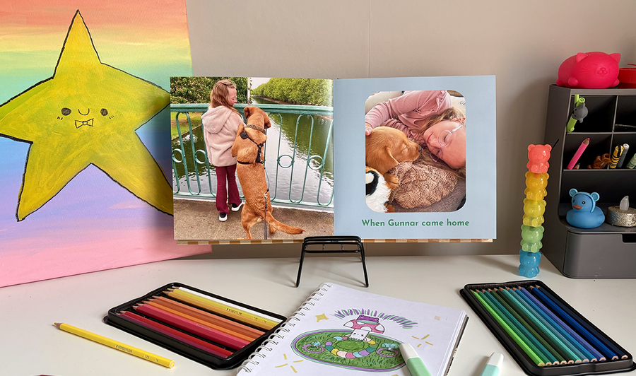 En åpen barnefotobok ligger på et barns skrivebord omgitt av fargestifter og kunstverk, med et bilde av en jente og hunden hennes på venstre side og et annet bilde med bildetekst på høyre side, som illustrerer fotoknudsen sine tips for å blande bilder og kunstverk i en familiefotobok.