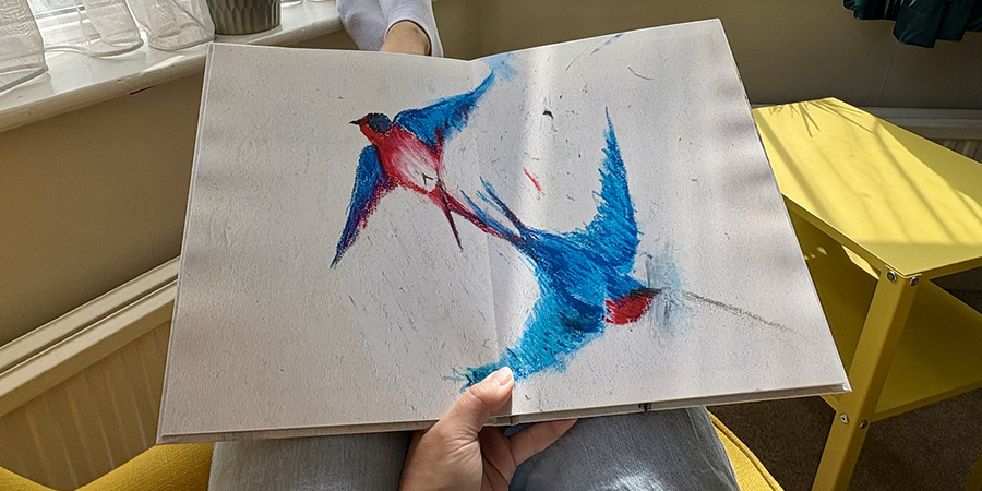 En åpen barnefotobok med et fullsides oppslag av en fugletegning ligger på skjørtet til en kvinne som sitter i en lenestol, som illustrerer fotoknudsen sine steg for å lage en familiefotobok av barnets tegninger.