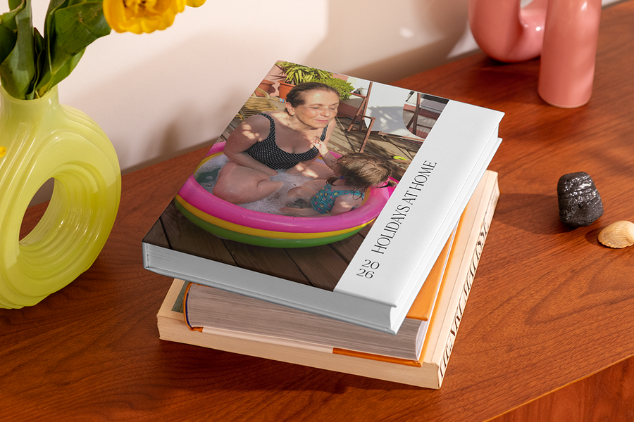 En stabel med fotobøker har på toppen en bok som viser en kvinne og et lite barn i en oppblåsbar plaskebasseng, med titelen 'Holidays at Home', som illustrerer fotoknudsen sine fotobok tittel ideer for staycation.