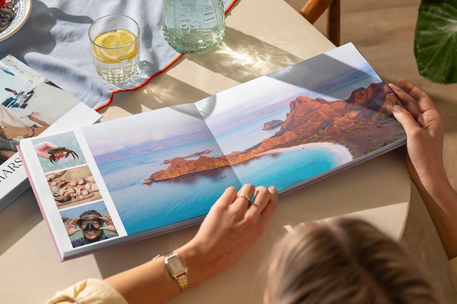 En åpen reisefotobok på et bord viser et dobbeltsidig oppslag med et vakkert strandlandskap sammen med tre sommerbilder, som illustrerer fotoknudsen sitt Travel Chapter-design fra den nye Summer Travel Series.