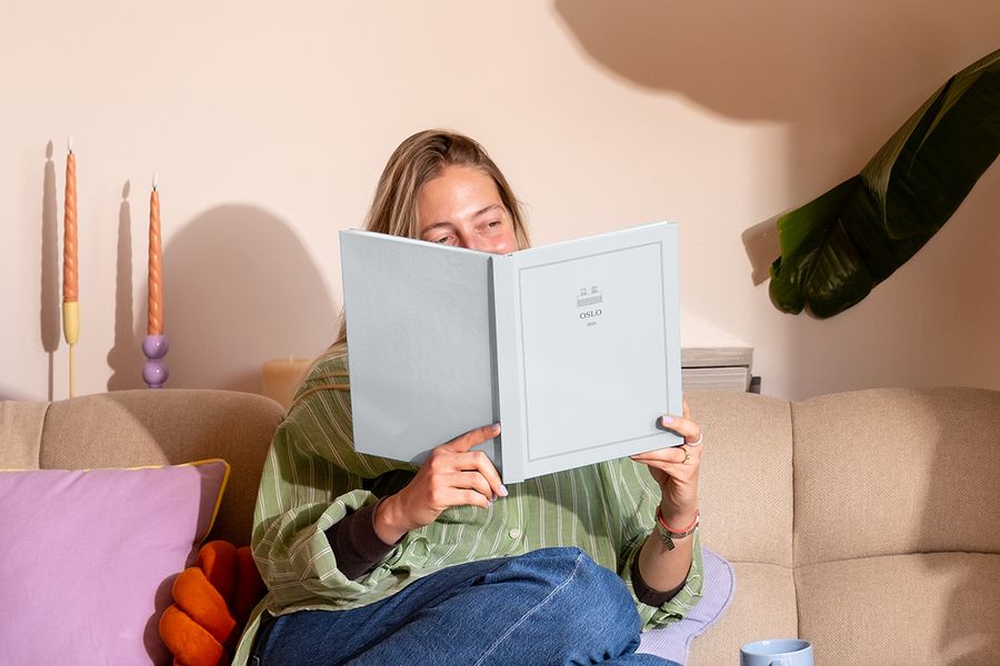 En kvinne sitter i sofaen ved siden av en fargerik pute og ser på en personlig reisefotobok med titelen 'Oslo', som viser fotoknudsen sine nye Summer Travel Series-design.