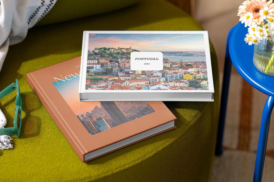 To personlige fotobøker ligger stablet på en krakk, den øverste med titelen 'Portugal' og et bilde av Portugal, og under den en bok med titelen 'New York' og et bilde av byen, som viser fotoknudsen sine personaliserbare Summer Travel Series-design.