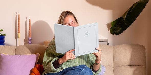 En kvinne sitter i sofaen ved siden av en fargerik pute og ser på en personlig reisefotobok med titelen 'Oslo', som viser fotoknudsen sine nye Summer Travel Series-design.