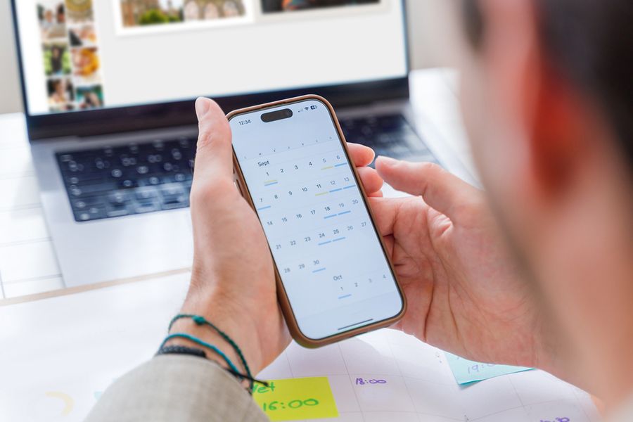 Noen ser på kalenderen på telefonen sin mens de i bakgrunnen jobber med en fotobok på laptopen, som illustrerer fotoknudsen sin enkle månedlige rutine for bildevedlikehold.