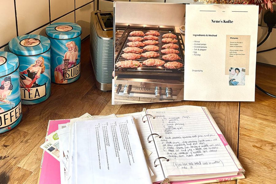 Sarahs åpne personlige kokebok viser en oppskrift på Nenes kofte, plassert foran en gammel mappe åpnet for å vise håndskrevne familieoppskrifter, begge liggende på en kjøkkenbenk, som illustrerer hvordan hun samlet generasjoners oppskrifter i en bok.