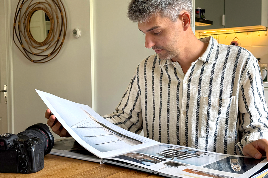 Charl van Rooy sitter på kjøkkenet sitt og ser ned på den åpne porteføljen sin, med kameraet ved siden av seg på bordet, som illustrerer fotoknudsen sin guide til å lage en fotografiportefølje som fotobok.