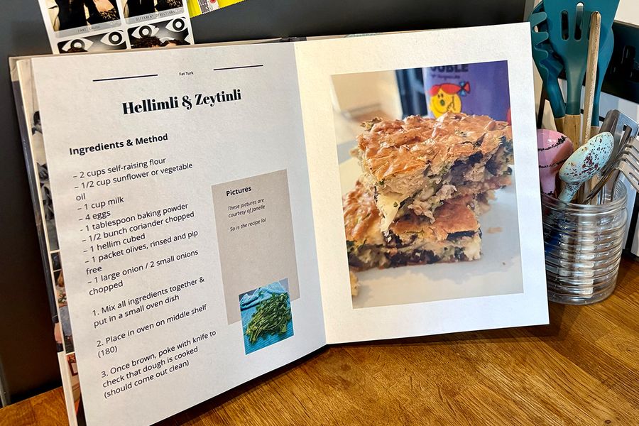 En åpen personlig kokebok ligger på et kjøkken, med en krukke med skjeer og kjøkkenredskaper i bakgrunnen, som illustrerer fotoknudsen sine tips til å velge hvilke oppskrifter som skal være med før du lager din egen kokebok.