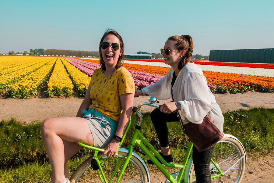 To kvinner sykler sammen, smiler og ler i et blomsterfylt felt, som illustrerer fotoknudsen sine idéer til hva man kan skrive i et bryllupsdagskort til venner.