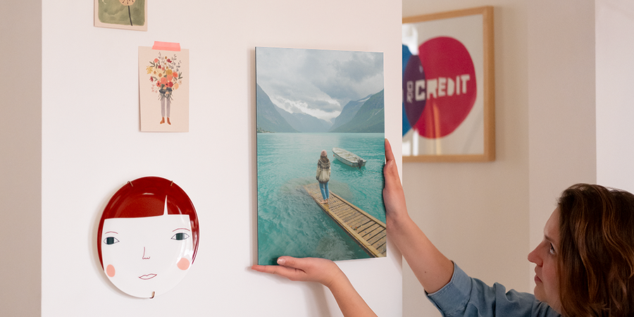 En kvinne setter opp personlig veggkunst på veggen sin, ved siden av en dekorativ tallerken og noen fotoutskrifter, som introduserer fotoknudsen sin trinn-for-trinn-guide til å henge opp veggkunst med opphengskit.