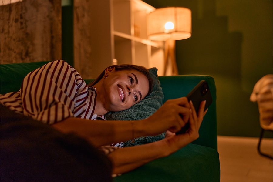 En kvinne ligger på en mørkegrønn sofa og ser på telefonen sin i en stue, opplyst av en lampe for lun belysning, som illustrerer fotoknudsen sine tips til hygge-belysning hjemme.