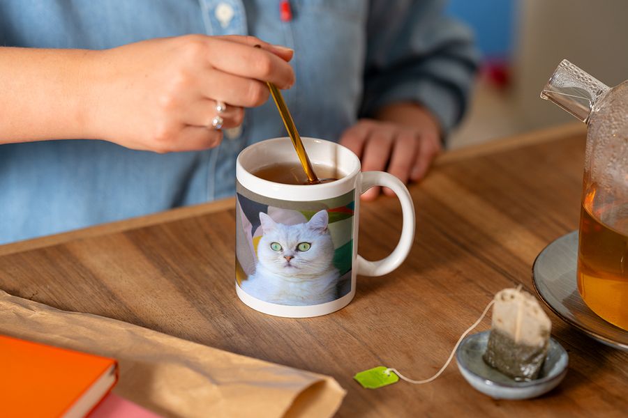 Noen rører med en skje i en personlig fotokopp med et bilde av en hvit katt mens de lager en kopp te, som illustrerer fotoknudsen sitt tips om å fullføre en hygge-lesekrok med en personlig kopp.
