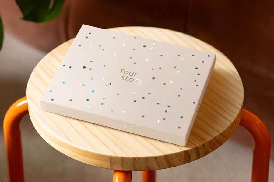 En fotoknudsen presentasjonsboks står på en trekrakk, dekorert med sølvfargede holografiske sirkler og teksten 'Your Story', som illustrerer det matte elfenbensfargede designet med et snev av holografisk glans.