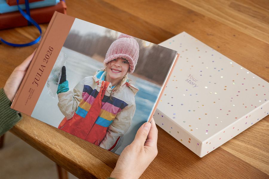 Noen holder en fotobok med titelen 'Julia', med en smilende jente på forsiden, med en presentasjons-gaveboks liggende rett bak på bordet, som illustrerer hvorfor en fotobokboks gjør en kreasjon ekstra spesiell.