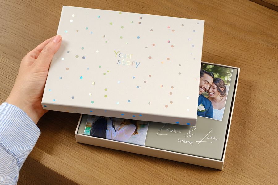 En bryllupsfotobok ligger i den nye presentasjonsboksen, med et bilde av paret og bryllupsdatoen som titel på forsiden, delvis skjult av det gapende lokket på gaveboksen, som illustrerer hvorfor en presentasjonsboks er verdt det for en premium bryllupsfotobok.