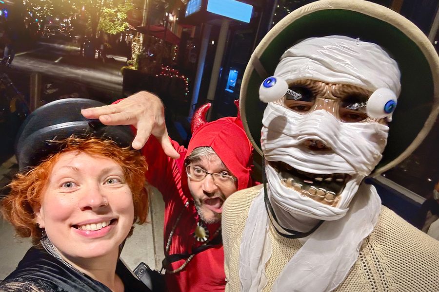 Tre voksne i Halloween-kostymer på gaten, en med skjelettmaske med utstående øyne, en utkledd som djevel og en kvinne med svart hatt, som illustrerer fotoknudsen sine Halloween-uttrykk for kostymer.