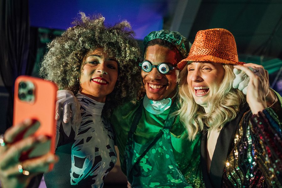 Tre voksne poserer for en selfie på en Halloween-fest, en kvinne i skjelettkostyme, en mann utkledd som skummel kirurg med morsomme briller og en kvinne med paljettbesatt hatt, som illustrerer fotoknudsen sine Halloween-uttrykk for grupper.