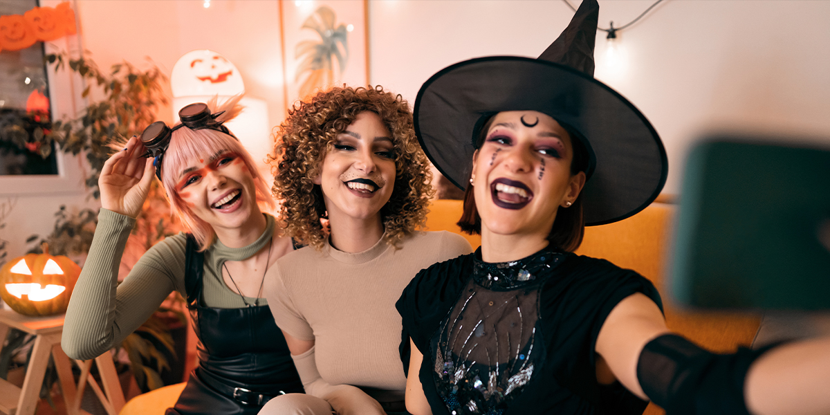 Tre kvinner i kostymer på en Halloween-fest, en heks, et dyr og en cyberpunk, poserer for en selfie på telefonen, som introduserer fotoknudsen sine 105 Halloween-sitater for Instagram.