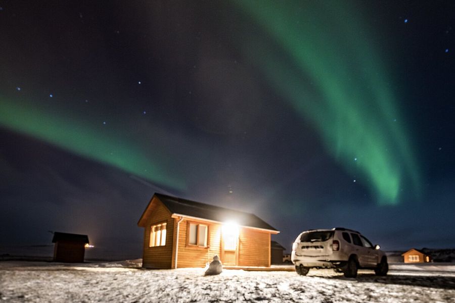 En hytte med lys som skinner ut, ved siden av en bil i et veldig snødekt landskap, med grønt nordlys på himmelen over, som illustrerer fotoknudsen sin forklaring på hva nordlys er og hva som forårsaker aurora borealis.