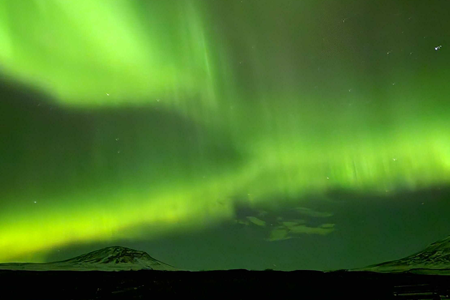 Snødekte fjell under en himmel som er helt grønn av nordlys, som illustrerer fotoknudsen sine tips om hvor man kan se nordlys uten å reise til Island.