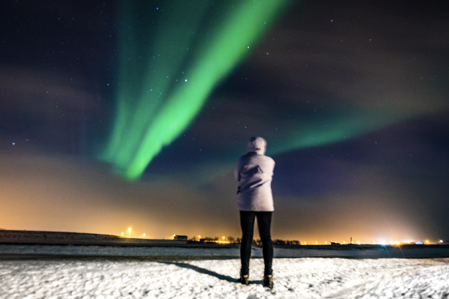 En person som står i et snødekt landskap og ser opp mot det grønne nordlyset på himmelen, som illustrerer fotoknudsen sine tips for å fotografere nordlys med mobil.