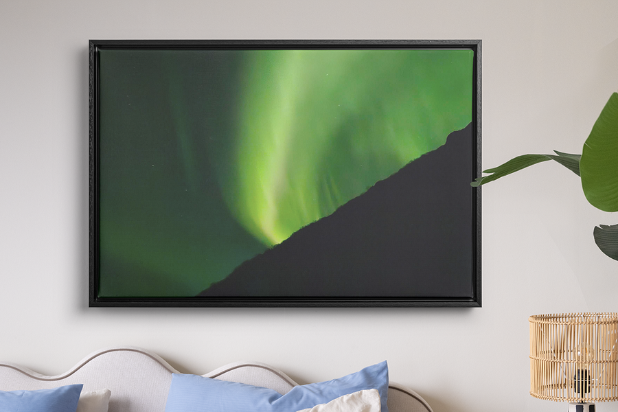 Et innrammet bilde av veldig grønt nordlys henger på veggen over en seng og en rafialampe, som illustrerer fotoknudsen sin veggkunst for å gjøre nordlysbildene dine til noe verdt å beholde.