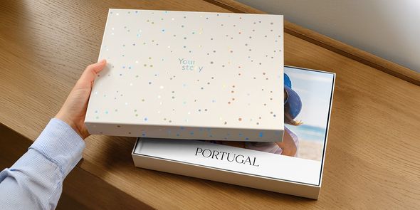 En fotoknudsen presentasjonsboks står med lokket på gløtt, og viser en personlig reisefotobok for Portugal inni, med forsiden delvis skjult av lokket, som introduserer fotoknudsen sin nye fotobok-gaveboks.