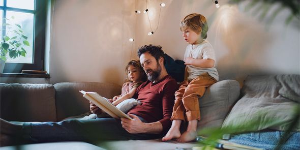 En far leser en bok for en liten gutt og en liten jente i sofaen, med varme lyslenker foran seg, som introduserer fotoknudsen sine tips til hyggelig høstinnredning i hygge-stil.