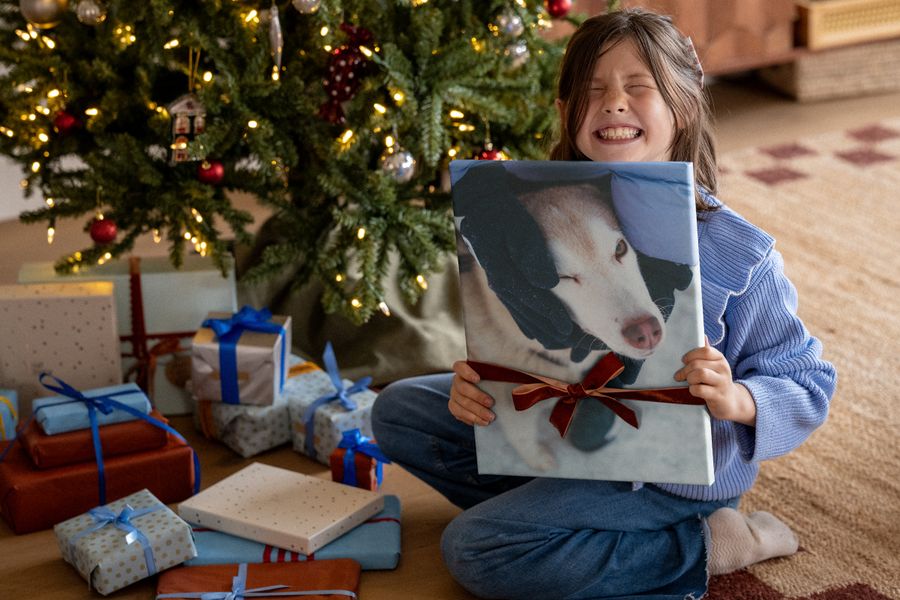 En jente som ler høyt i en blå genser holder opp et lerretsbilde av en hund med et rødt bånd rundt, foran et juletre, til illustrasjon av fotoknudsen sine tips om veggkunst som Black Friday-julegave.