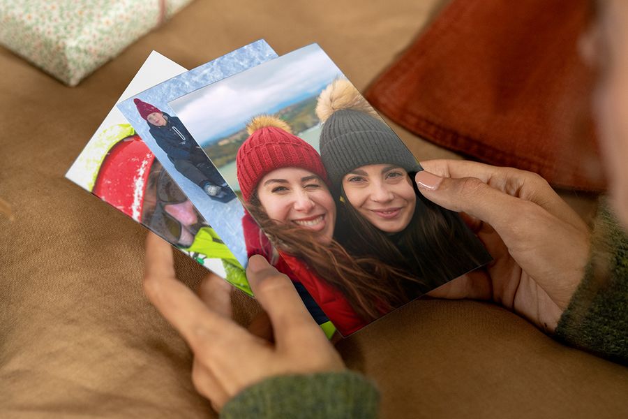 Person som holder fotoutskrifter i hendene, der den fremste viser to smilende kvinner i luer og kåper, som illustrerer fotoknudsen sin guide til å finne de riktige bildene til en personlig fotogave.