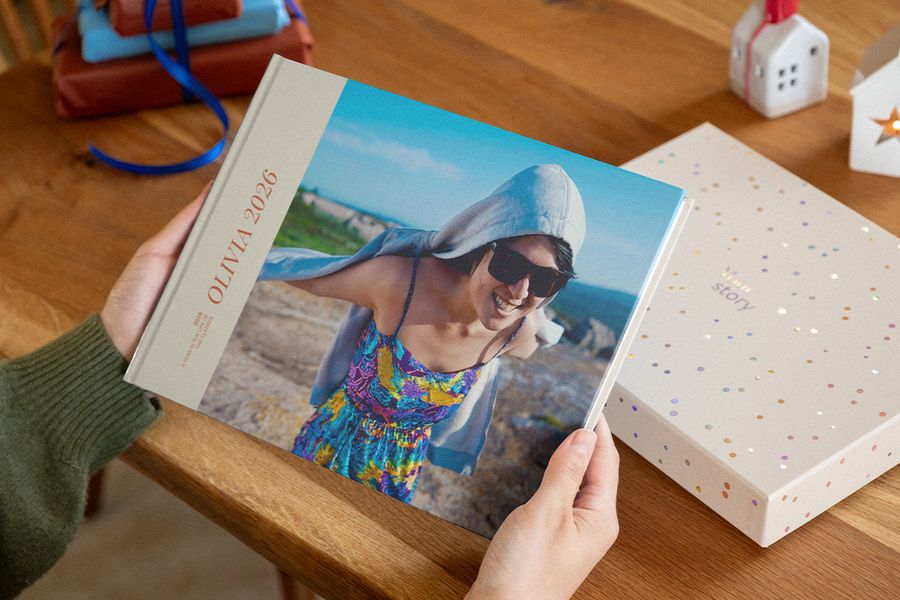 To hender holder en fotobok med 2026 på forsiden, der en smilende kvinne i solbriller er fotografert på en steinete kystlinje, ved siden av en presentasjonsboks med konfetti-prikker, til illustrasjon av fotoknudsen sine personlige fotobøker.