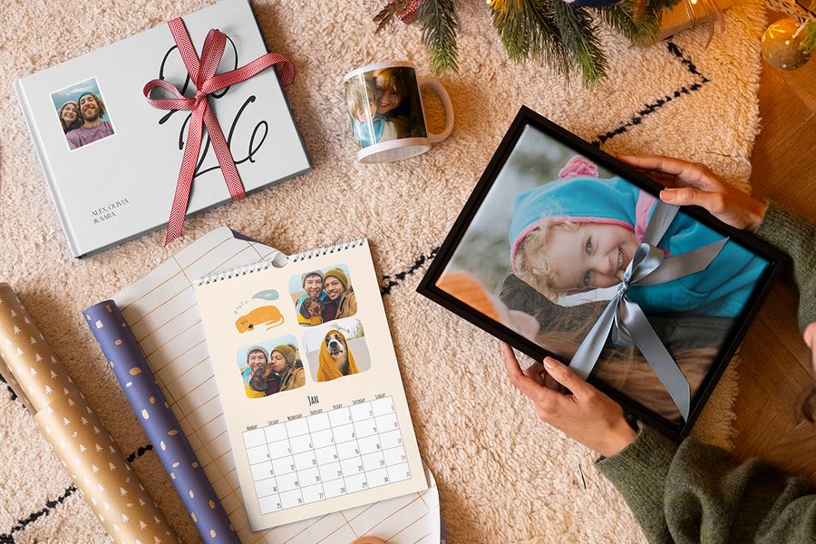 Utvalg av personlige gaver fra fotoknudsen, med veggkunst av en ung jente med et blått bånd rundt, et krus med en mor og et barn, en personlig fotokalender og en 2026-fotobok med et rødt bånd rundt, ved siden av gavepapir og med juletregreiner i bakgrunnen, som illustrerer fotoknudsen sin guide til å fange de riktige minnene i en personlig julegave.
