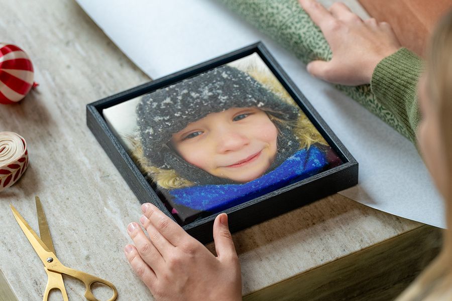 Innrammet lerret av en ung jente i vinterklær i snøen, med en kvinnes hender klare til å pakke det inn, ved siden av gavepapir, saks og bånd, som illustrerer fotoknudsen sin guide til å velge en personlig fotogave som passer til stilen til mottakeren.