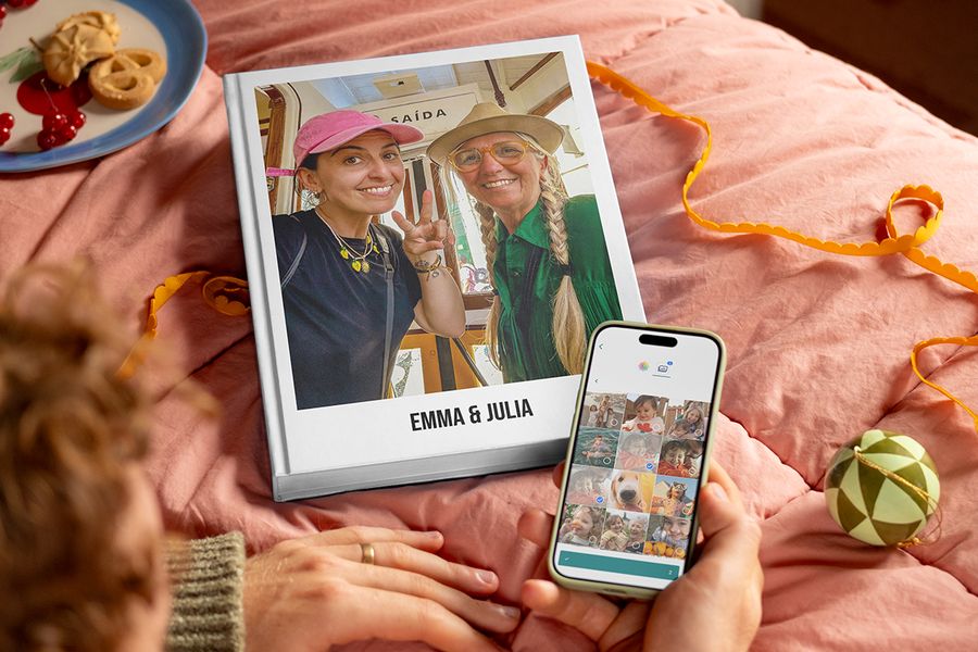 Personlig fotobok med tittelen “Emma and Julia” med en mor og hennes voksne datter på forsiden, ved siden av en manns hender som holder en telefon for å vise hvor enkelt det er å velge bilder og lage en fotobok fra fotoknudsen, som illustrerer fotoknudsen sin guide til å samle bilder fra familie og venner til en personlig gave.