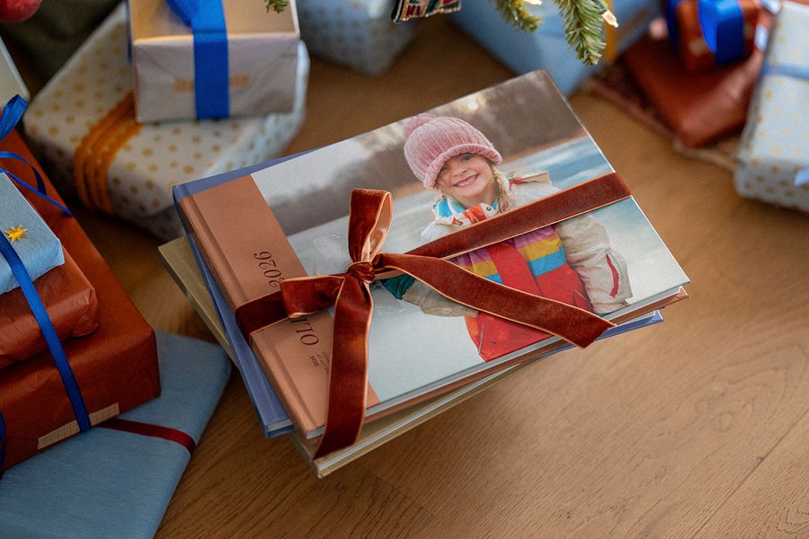 En stabel fotobøker bundet sammen med et fløyelsbånd, der den øverste boken viser en smilende jente i rosa lue og 2026 på ryggen, blant innpakkede julegaver, til illustrasjon av fotoknudsen sin fotobok for året.